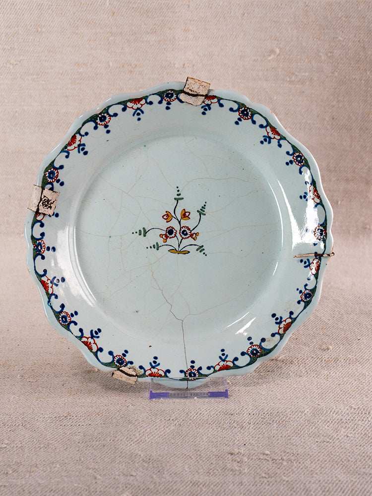 Antique French faience plate from Moustiers with scalloped edges and polychrome floral design, upright on neutral fabric.