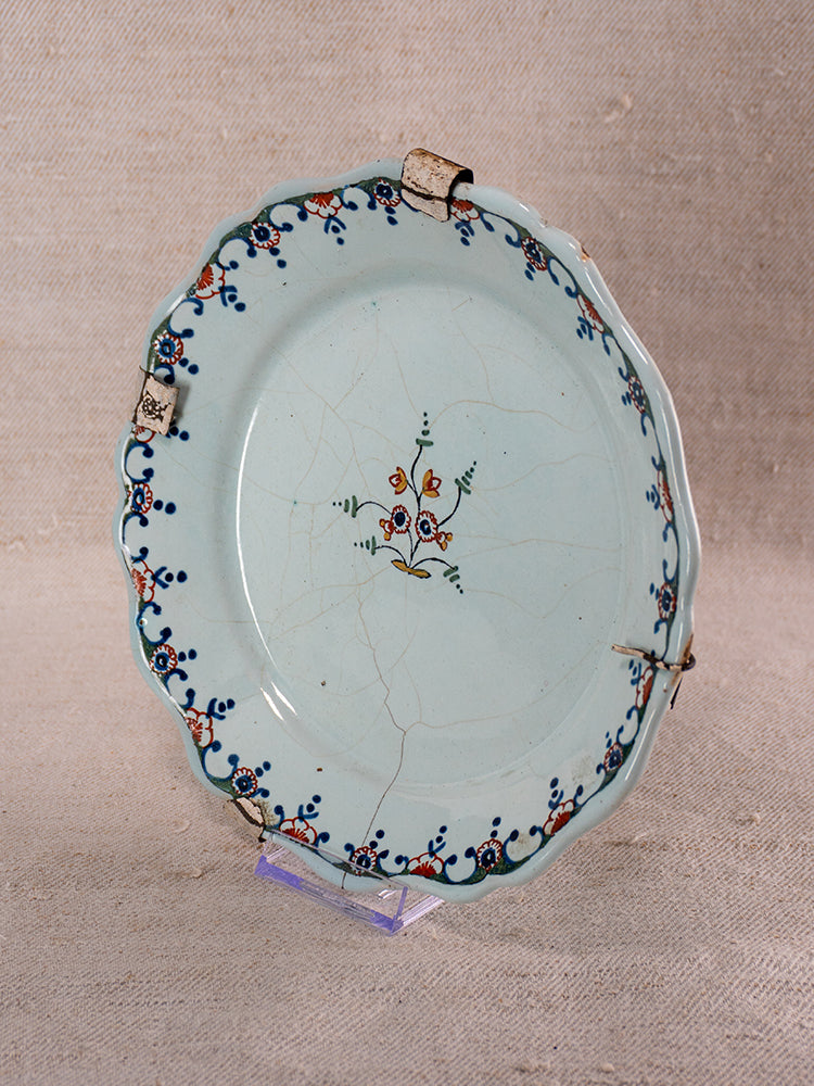 18th century Moustiers French faience plate, polychrome floral design, scalloped edge, metal-clipped repairs, on fabric background.