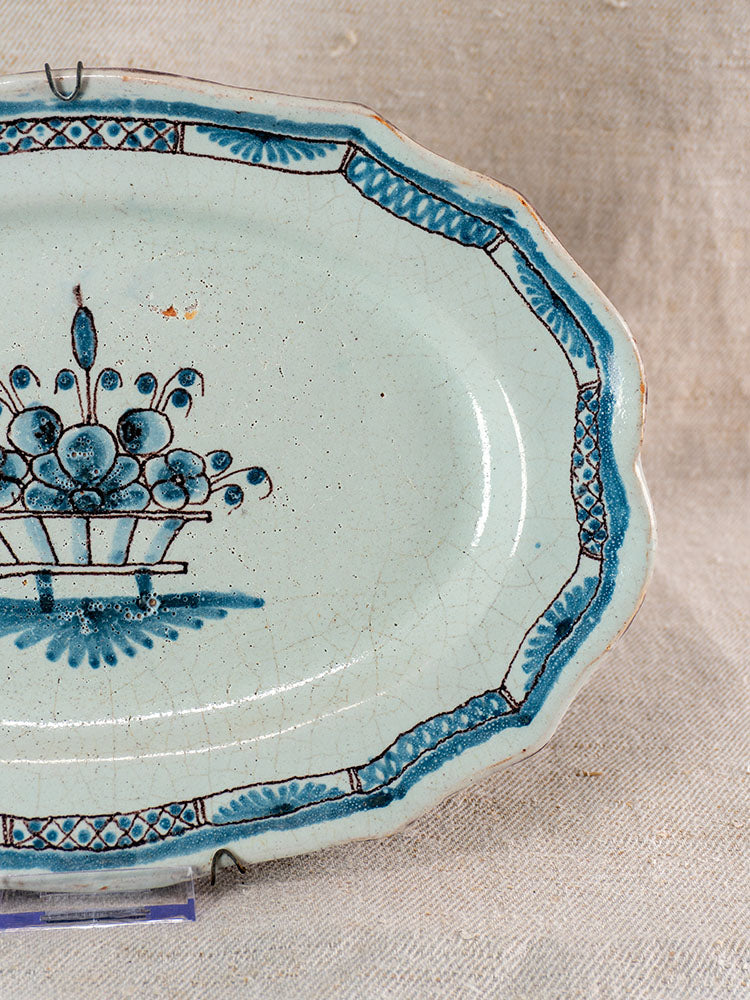 Antique French faience platter in Rouen Cul Noir style, blue floral basket center, ornate rim, beige textured fabric backdrop.