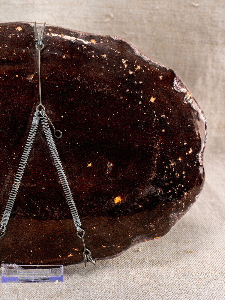 Antique French Rouen faience oval Cul Noir platter, dark brown, on metal display stand with burlap backdrop.