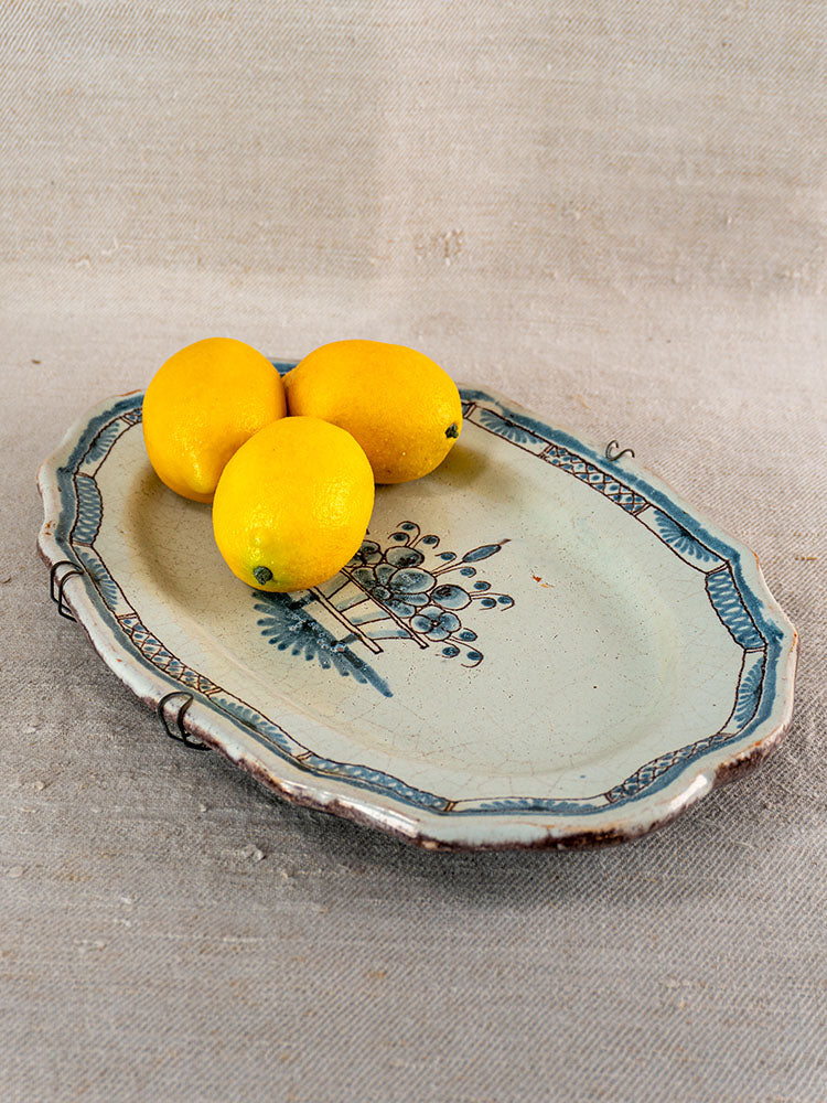 Antique French faience platter, Rouen tradition, blue and green motif, edge cracks, metal repairs, on textured beige fabric.