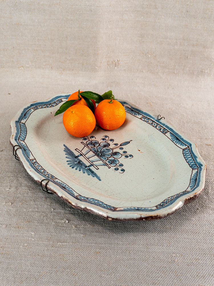 Antique French faience platter, Rouen Tradition Cul Noir style, blue floral motif, rustic edge, with oranges on beige fabric.