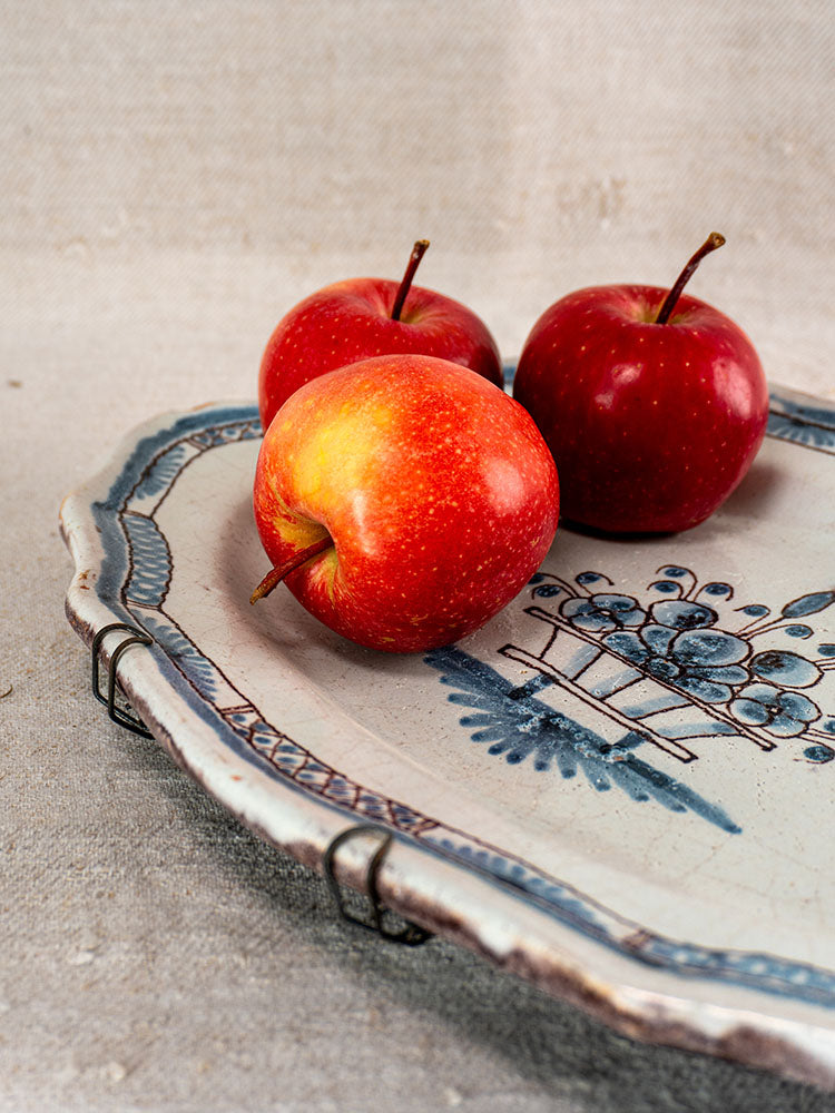 Antique French faience platter, Rouen Cul Noir style with blue floral design, displayed with red apples on textured fabric.