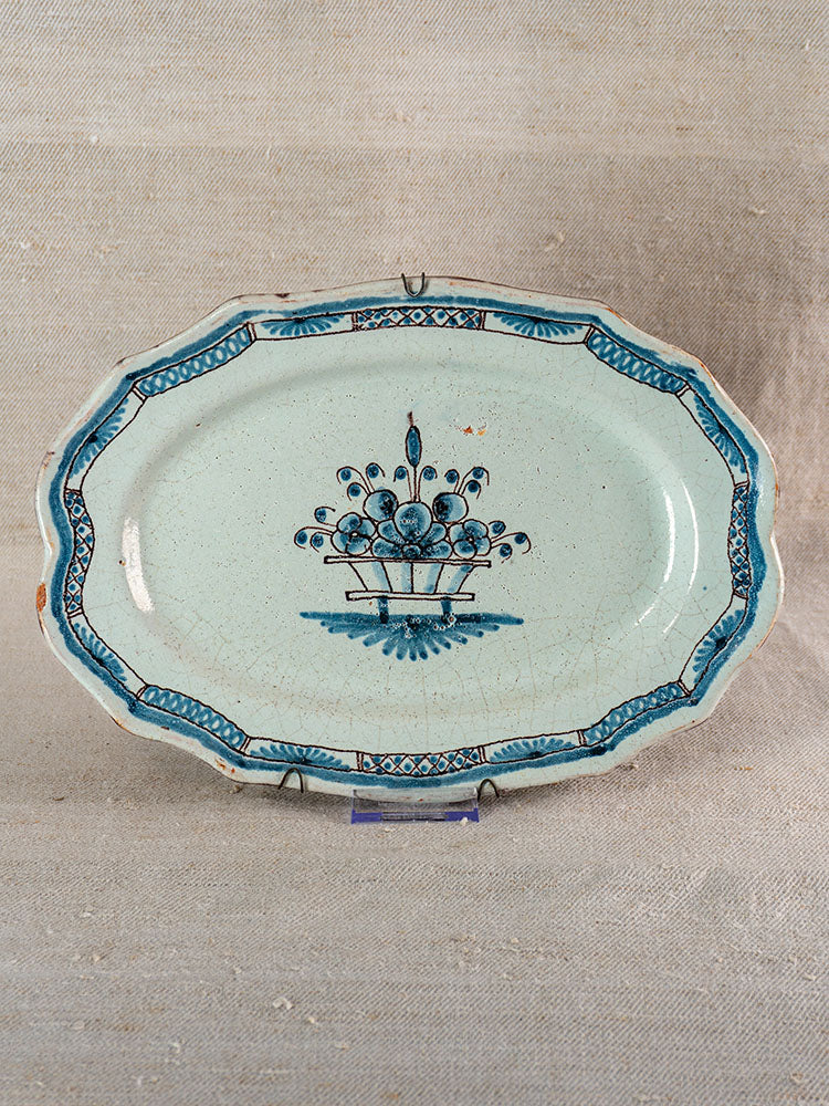 Antique French faience platter with scalloped edges, blue floral basket motif, on a neutral textured fabric background.
