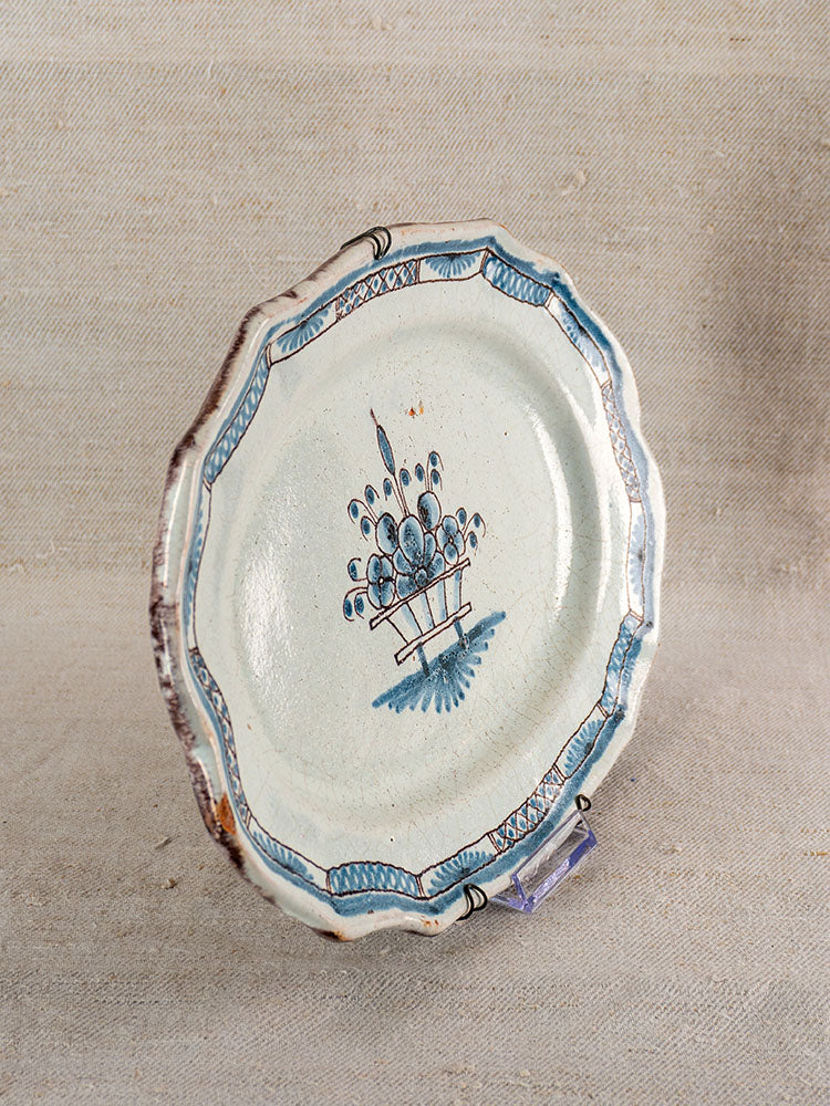 Antique French faience platter with scalloped edges, blue-brown floral center, and patterned rim on neutral fabric background.