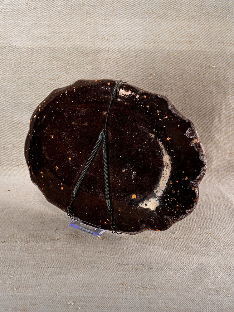 Antique French faience platter in Rouen Cul Noir style, dark brown speckled glaze, on a metal stand with beige fabric backdrop.