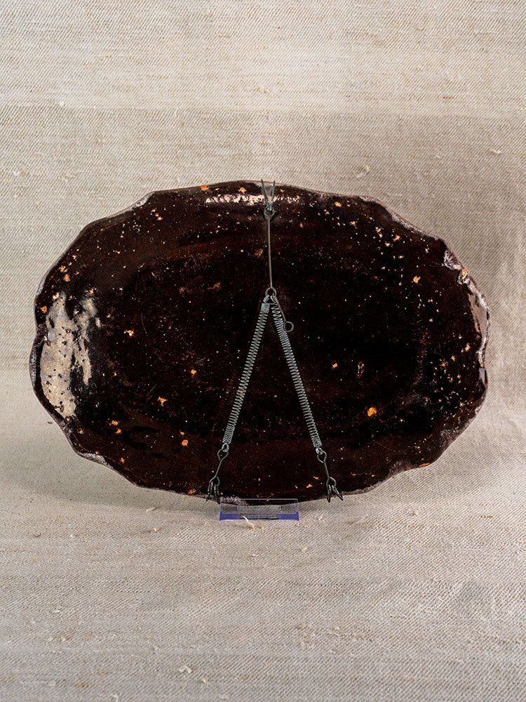 Antique French faience Rouen-style Cul Noir platter, dark brown, oval with uneven edge, on metal stand, beige textile backdrop.