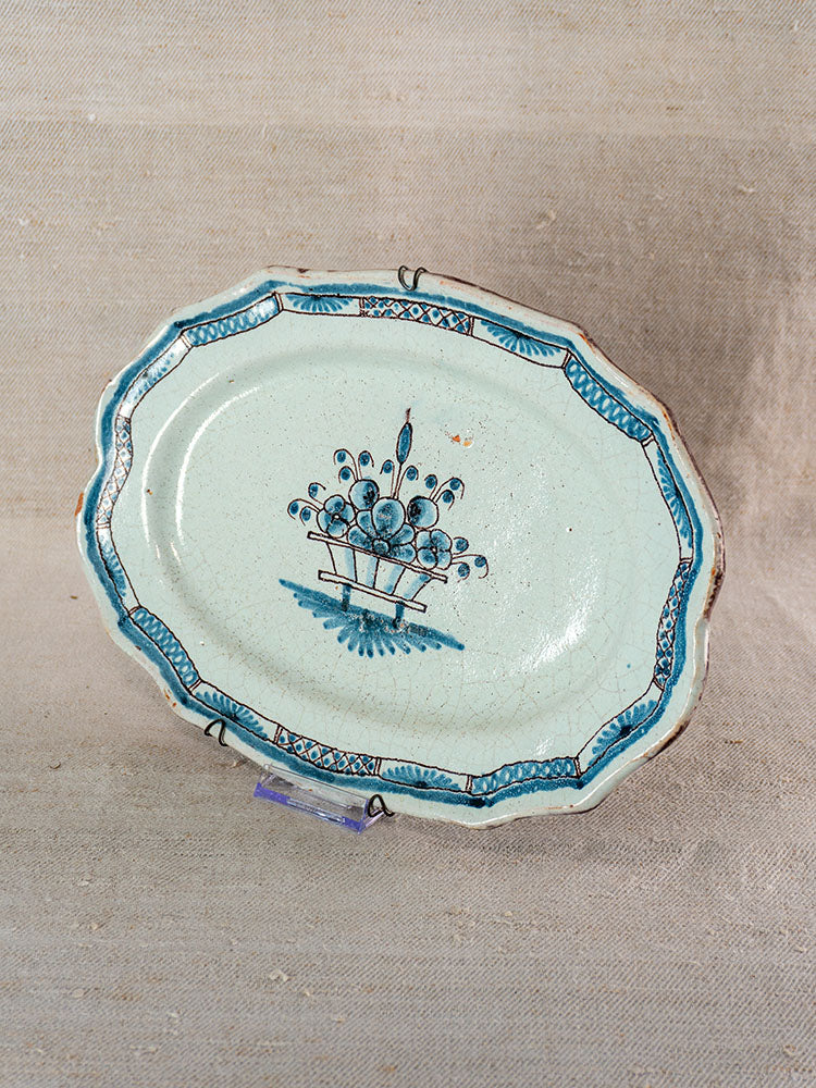 Oval antique French faience platter with light blue glaze, scalloped edge, and blue floral basket, on stand against neutral fabric.