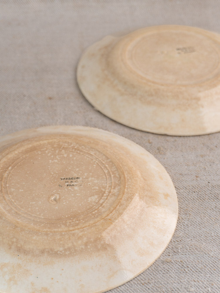 Antique French faience plates, HBCM Creil Montereau Trebeurden, upside down on beige textured fabric, aged bases visible.