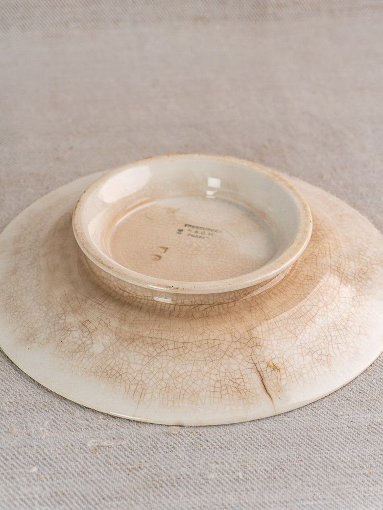 Antique French faience plate, HBCM Creil Montereau Trebeurden pattern, with crackle glaze, on neutral fabric beneath upside-down beige bowl.