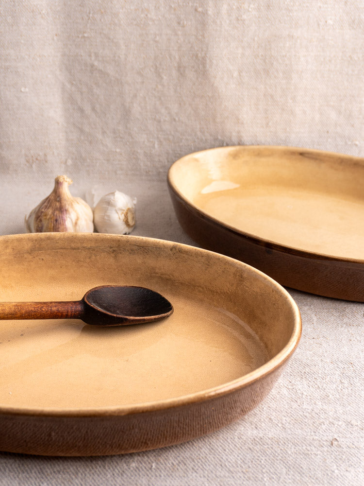 Pair of vintage French La Bourguignonne beige stoneware gratin dishes on textured cloth, garlic bulbs behind.