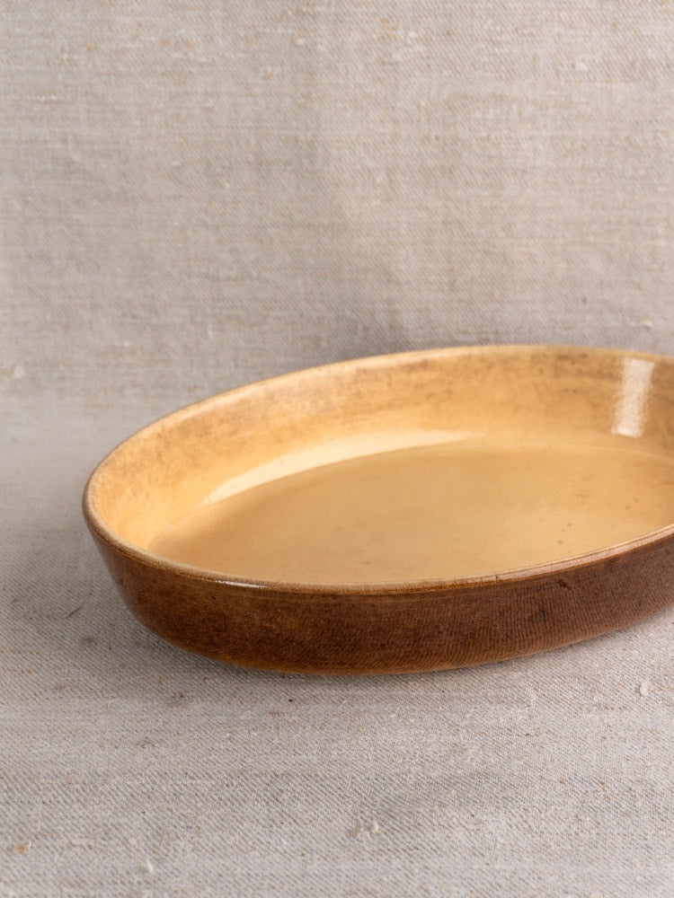 Shallow round brown ceramic La Bourguignonne gratin dish with glossy finish on beige fabric.