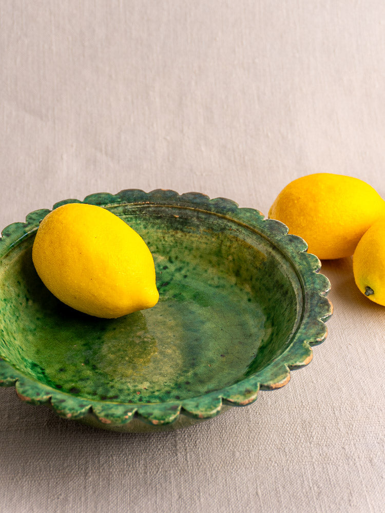 Antique French earthenware bowl with Provençal green glaze, filled with a lemon, on light fabric with two lemons beside.