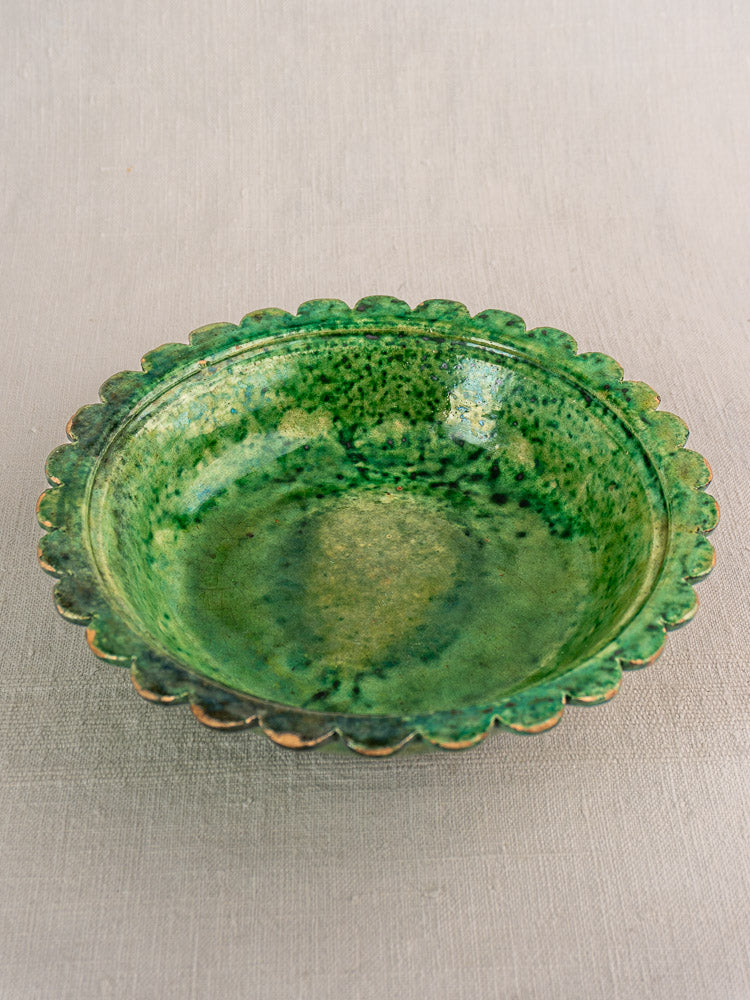 Antique French earthenware bowl with Provençal green glaze and scalloped edge on neutral fabric background.