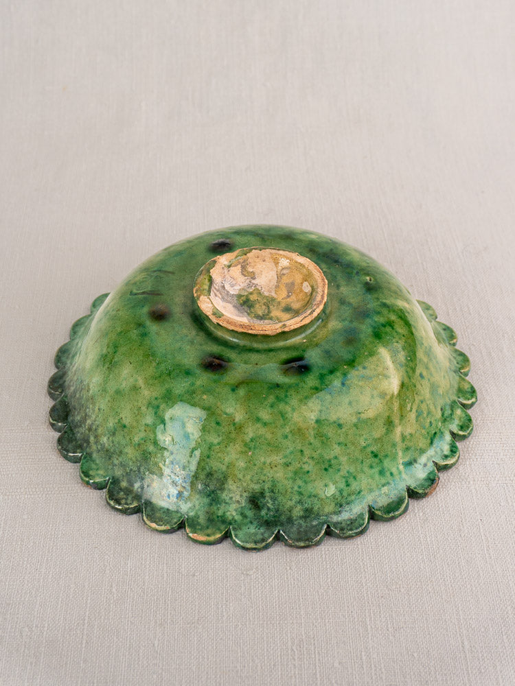 Antique French earthenware bowl with mottled Provençal green glaze, scalloped rim, unglazed foot on textured surface.