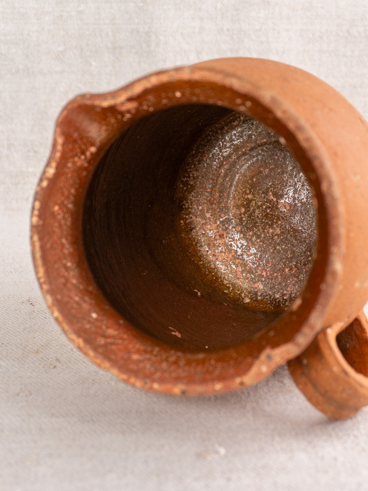 Antique French terracotta pitcher with pinched spout, top-down view on light fabric, showing textured, weathered interior.