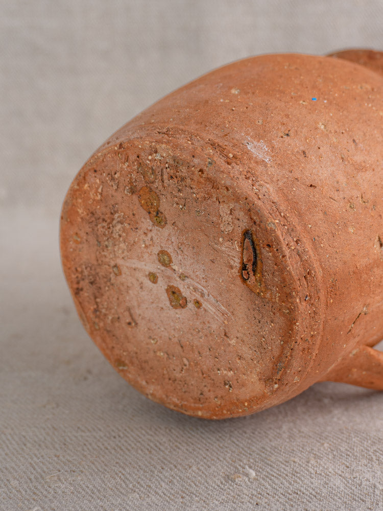Antique French terracotta pitcher base with pinched spout, textured surface and marks, on light fabric background.