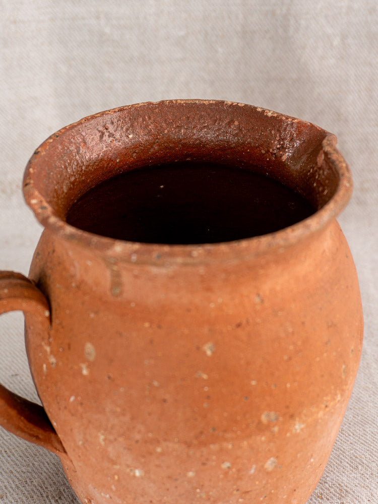 Antique French terracotta pitcher with pinched pouring lip and handle, rustic speckled finish, on light fabric.