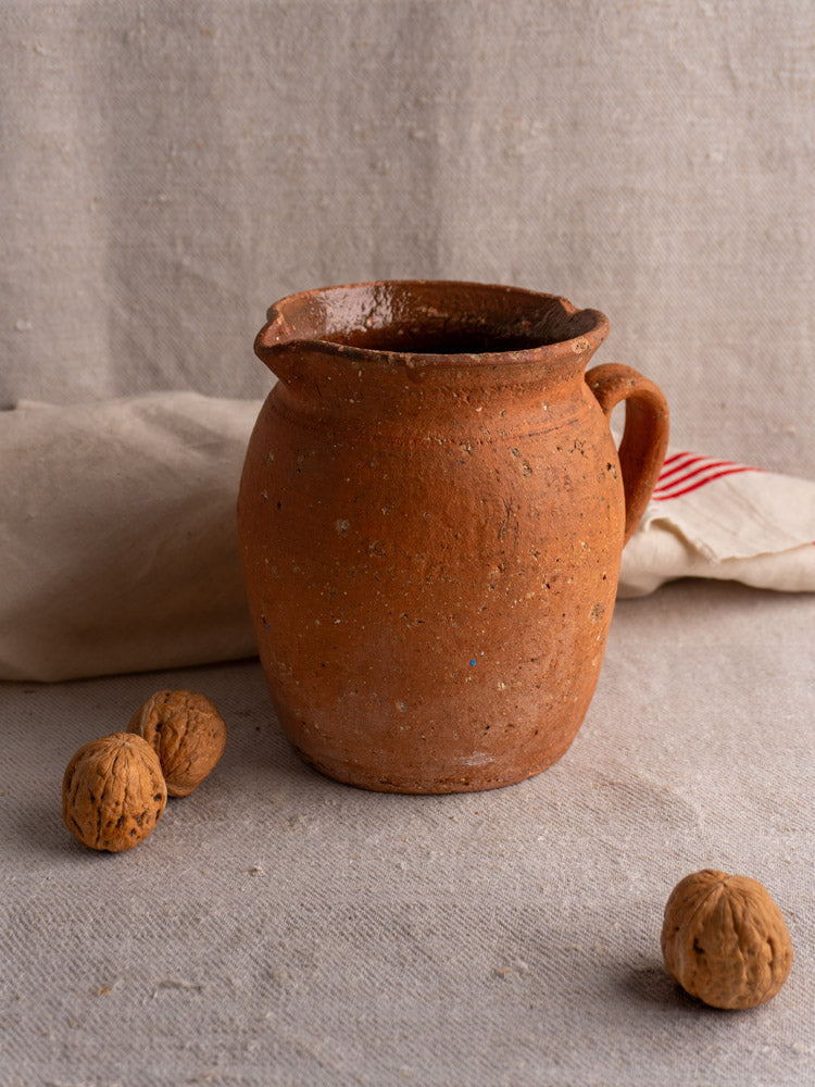Antique French terracotta pitcher with pinched spout, on beige fabric with three walnuts and folded cream cloth in background.