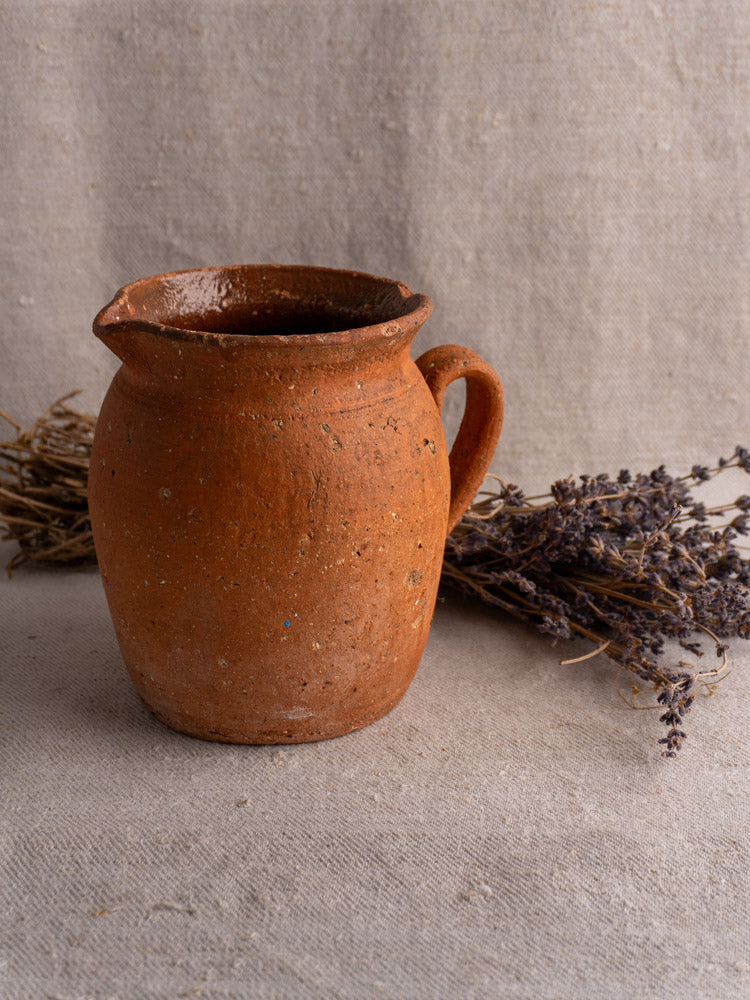 Antique French terracotta pitcher with pinched lip, mid to late 19th century, on textured beige fabric with dried lavender.
