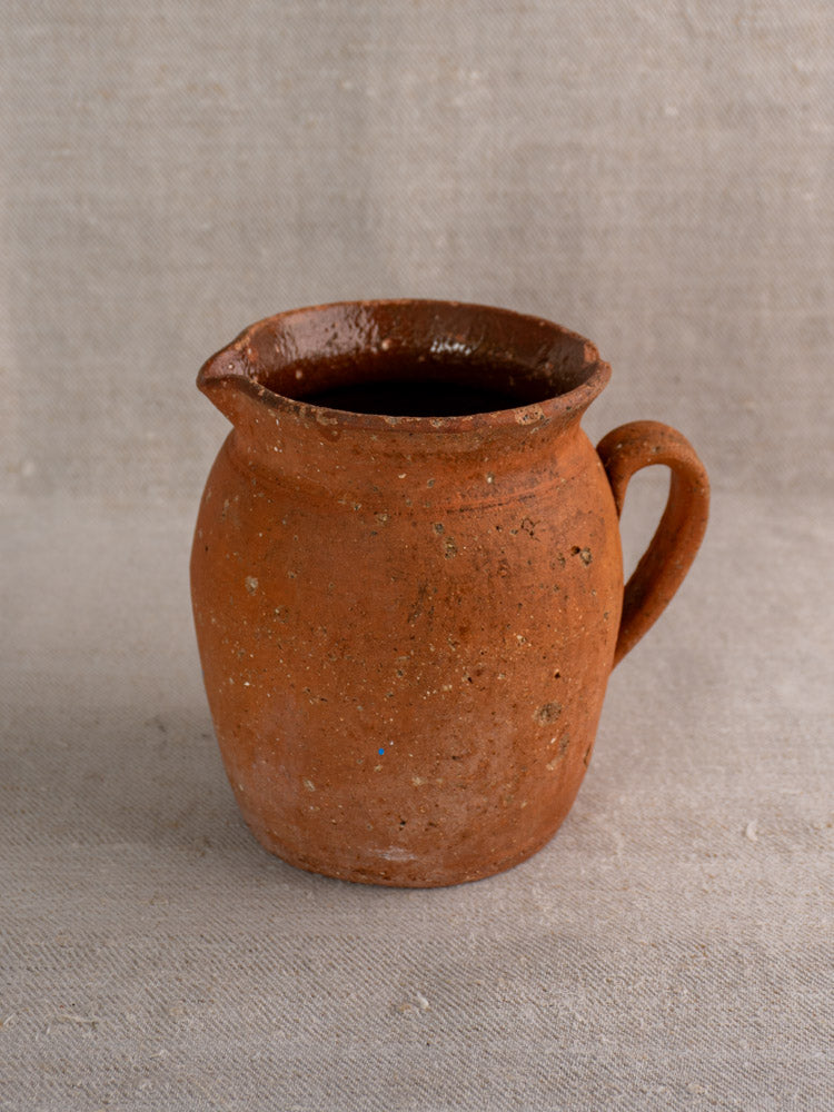 Antique French terracotta pitcher, mid-late 19th century, pinched lip and handle, reddish-brown speckled finish on beige fabric.