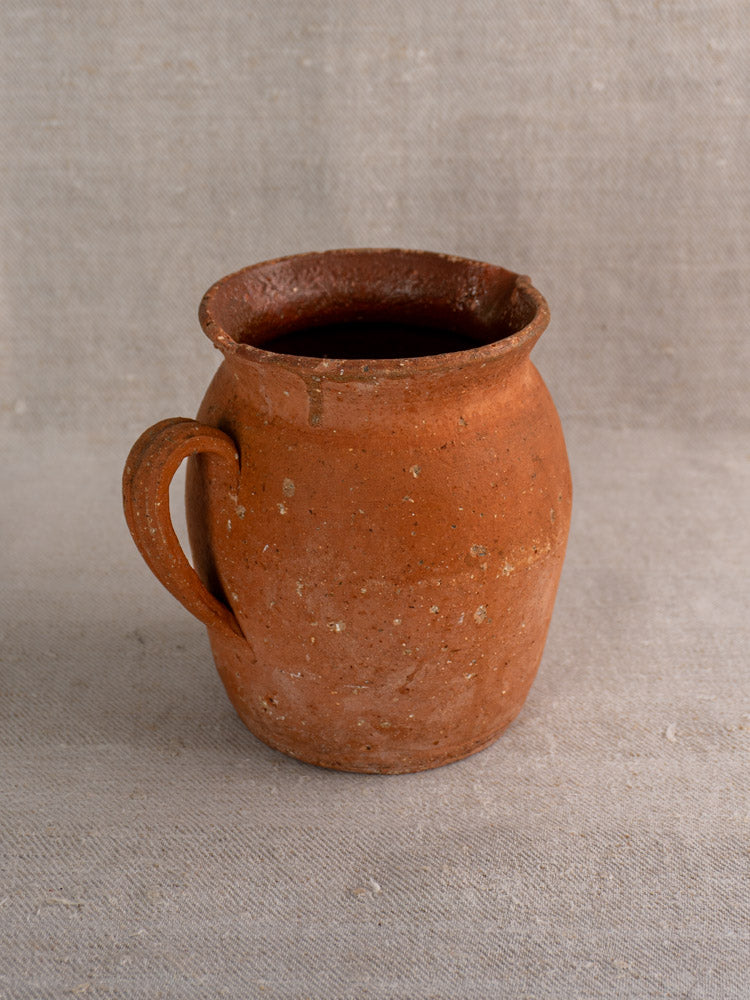 Antique French terracotta pitcher with pinched lip and handle on textured beige fabric, mid to late 19th century.