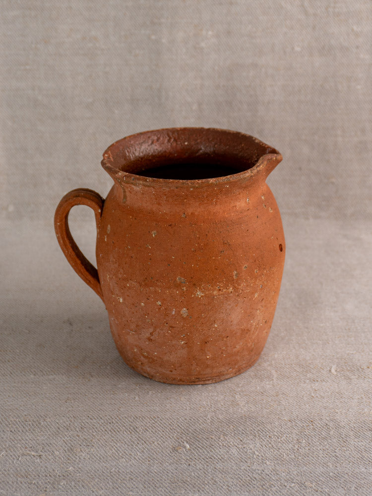 Antique French terracotta pitcher with pinched spout, handle, and weathered finish on textured fabric.