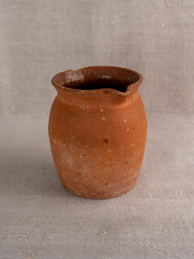 Antique French terracotta pitcher with pinched lip, reddish-brown finish, on textured beige fabric background.