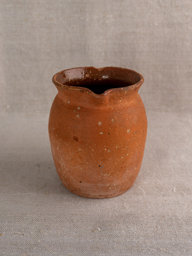 Antique French terracotta pitcher with pinched lip and rustic brown finish, on textured beige fabric background.