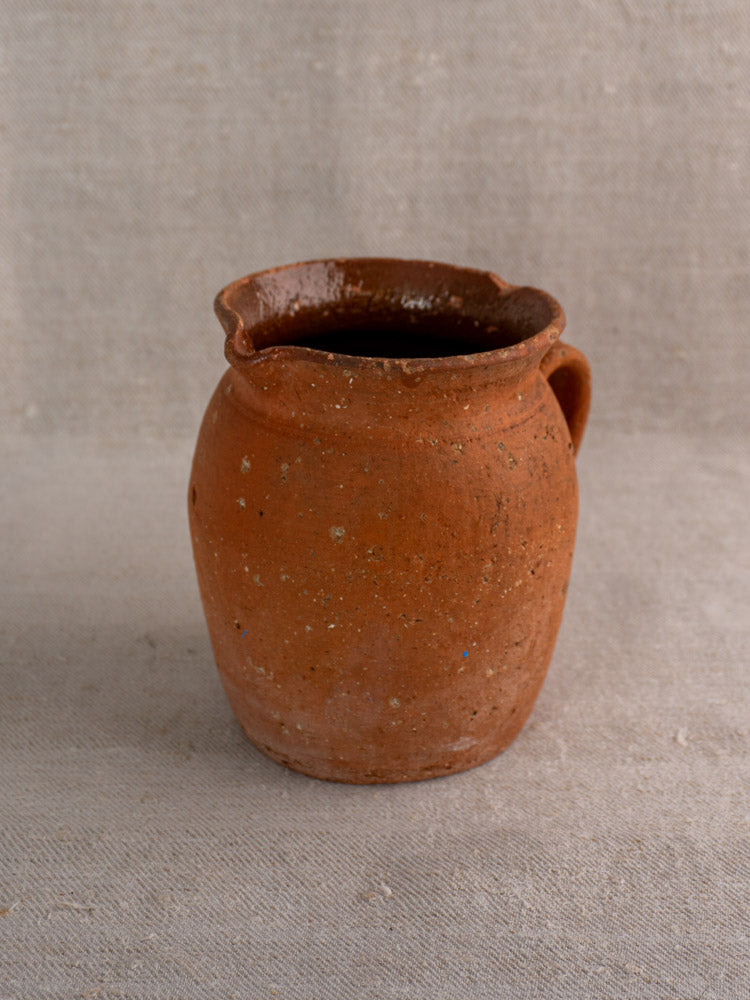 Antique French terracotta pitcher with pinched pouring lip and handle on textured beige cloth, rustic country pottery style.
