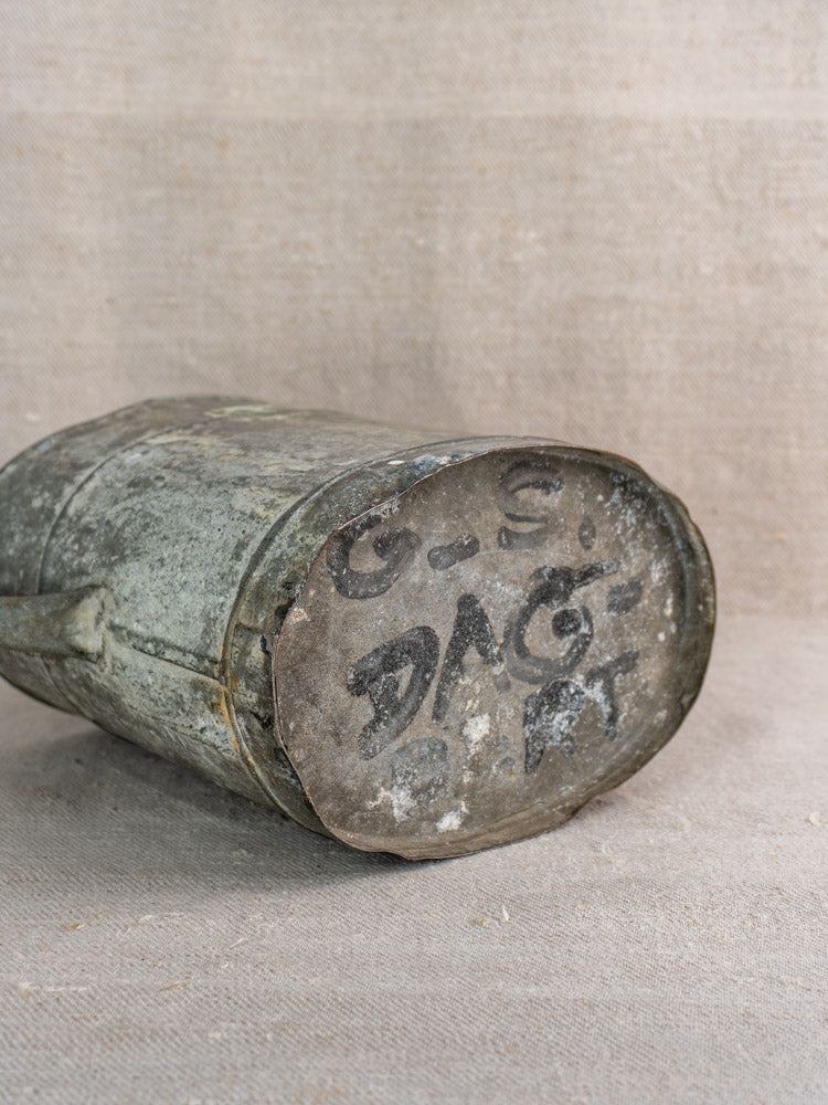 Antique French zinc watering can on textured fabric, showing worn base with faded black handwritten letters and numbers.