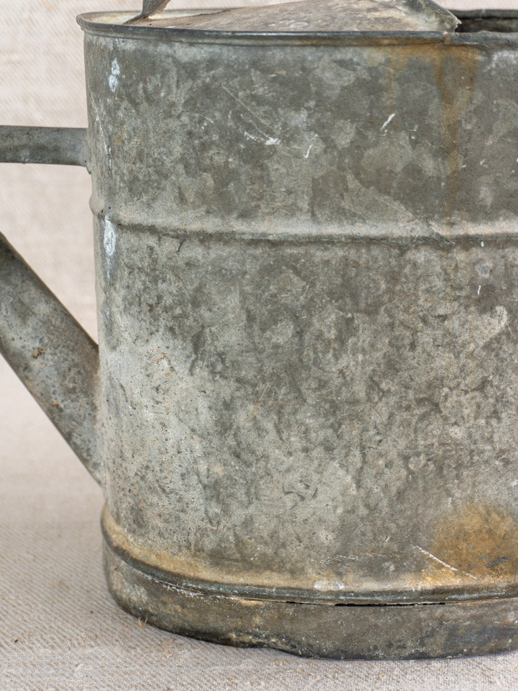 Antique French zinc watering can with handle, spout, and aged patina on textured fabric background.