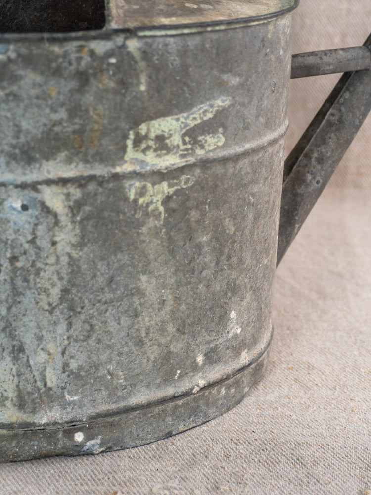 Antique French zinc watering can in galvanized sheet zinc, faint white mark on side, on textured beige fabric.