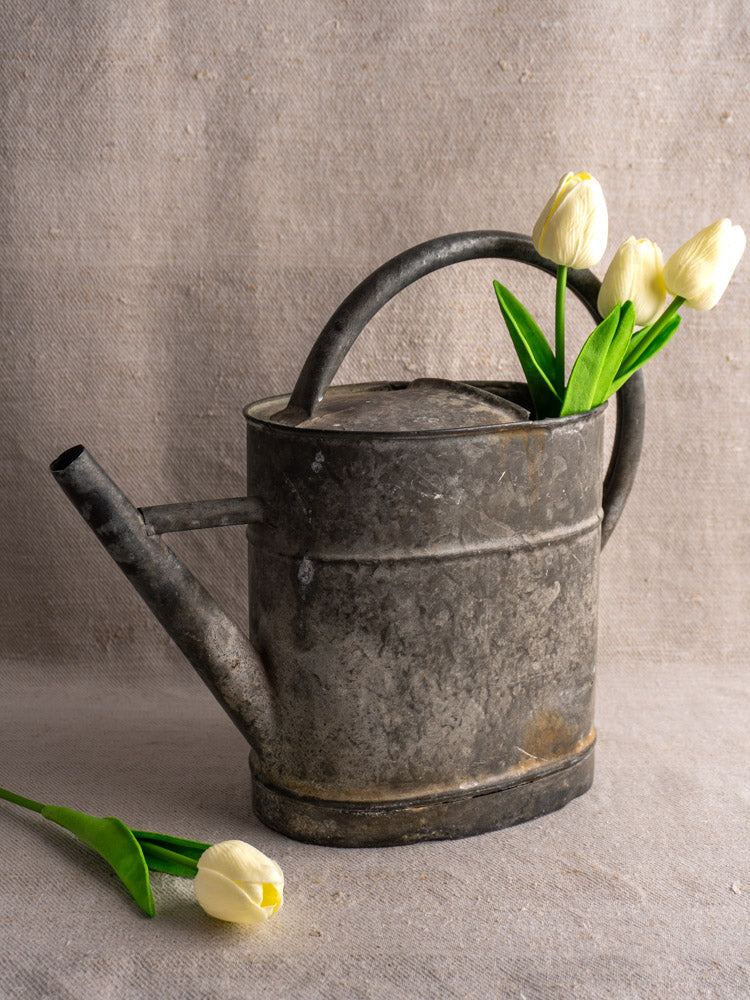 Antique French zinc watering can (late 19th–early 20th c.) with white tulips on textured beige fabric.