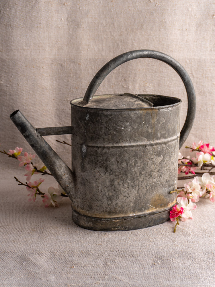 Antique French zinc watering can with curved handle and spout, set on beige fabric with pink and white blossoms.