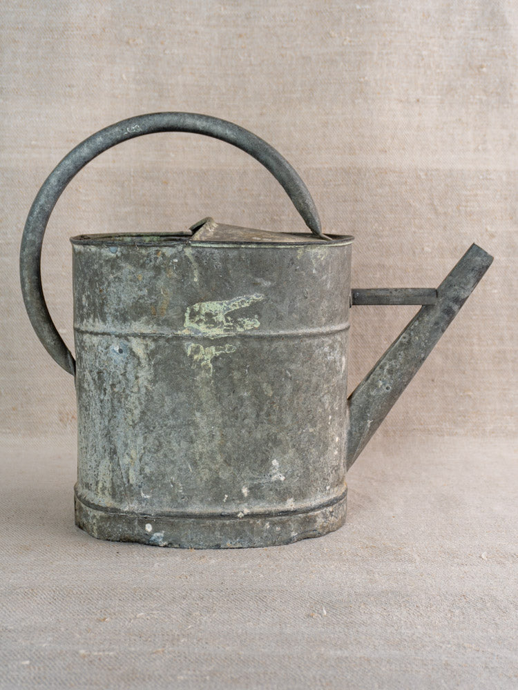 Antique French zinc watering can, late 19th–early 20th century, large handle, angled spout, aged patina on neutral fabric.