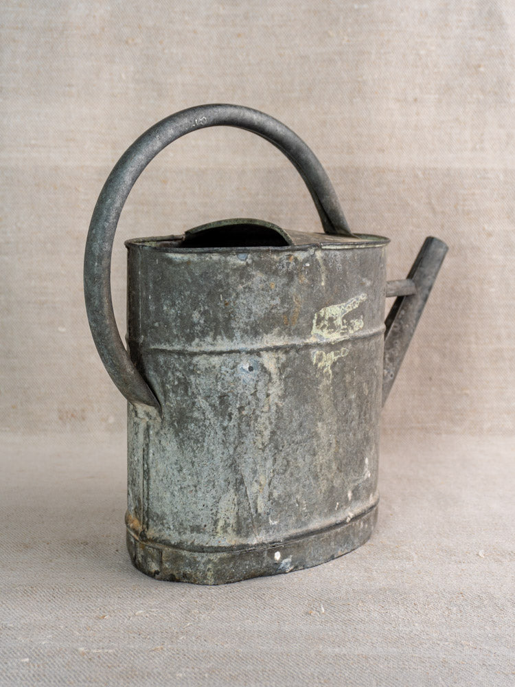 Antique French zinc watering can with curved handle and spout, showing rust and wear on a neutral textured surface.