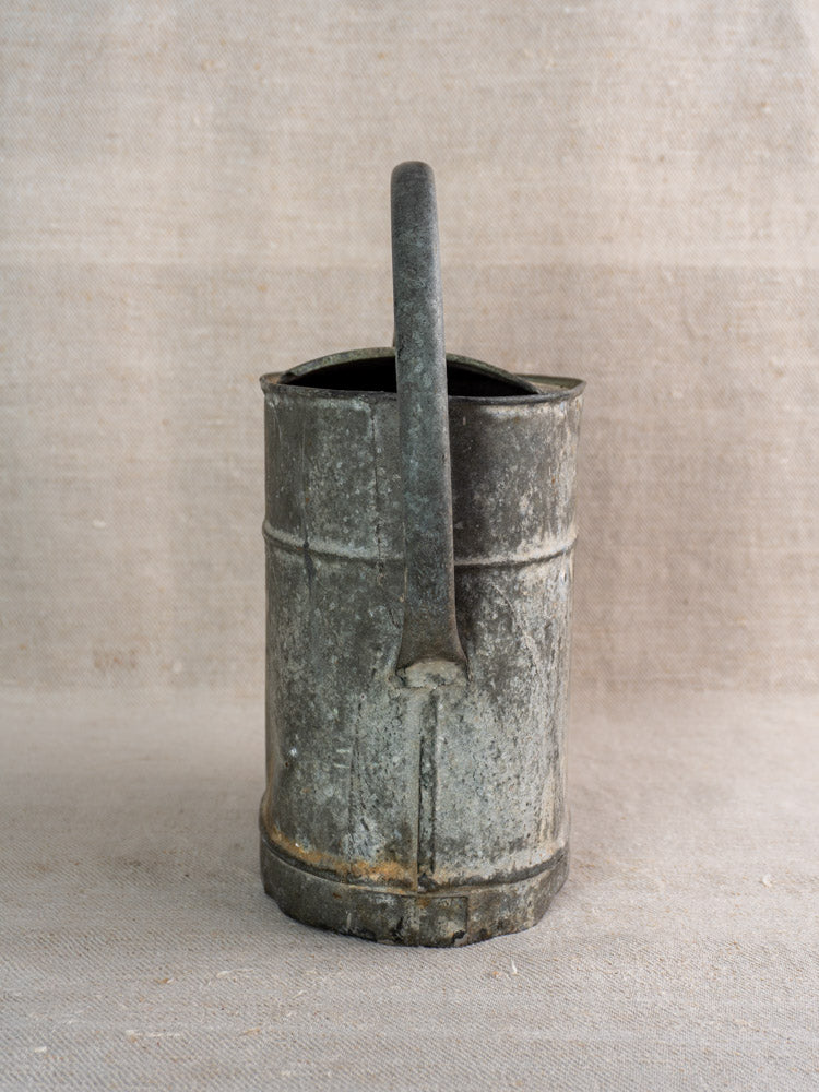 Antique French galvanized zinc watering can with curved handle and rustic patina on neutral fabric backdrop.
