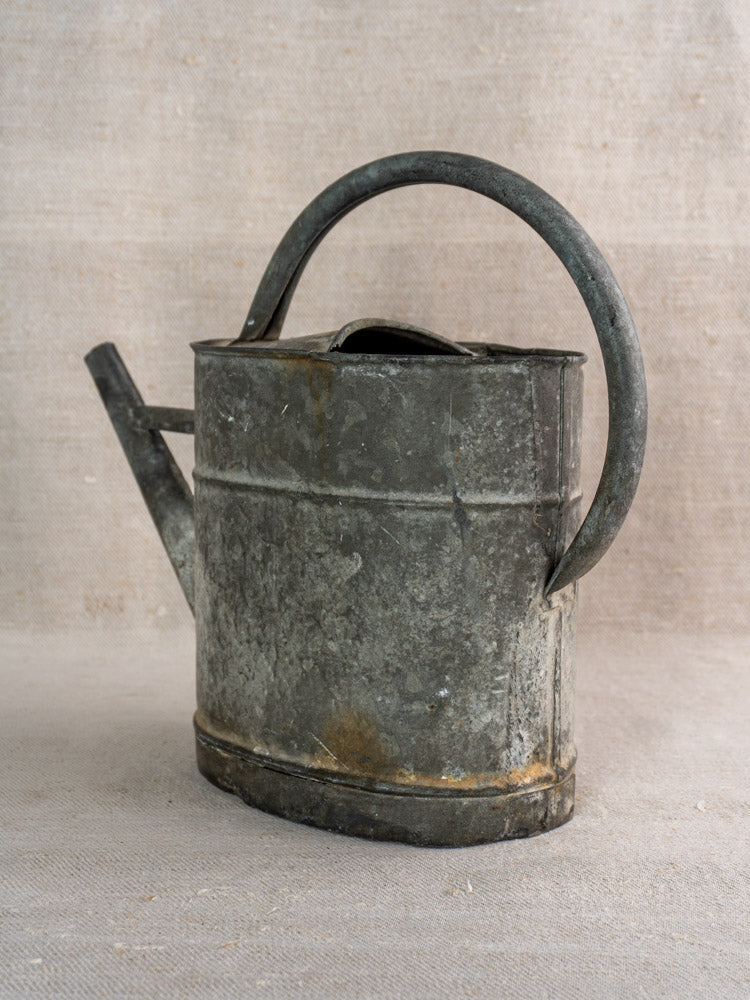 Antique French zinc watering can with curved handle and short spout on textured beige fabric background.