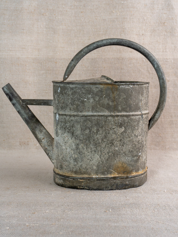 Antique French zinc watering can, late 19th–early 20th century, on neutral fabric with a beige background.