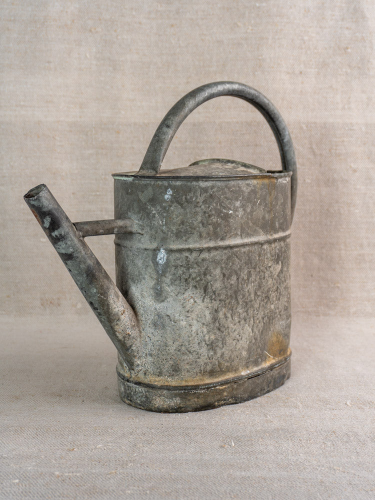 Antique French zinc watering can with curved handle and spout, displayed on neutral fabric against a beige background.