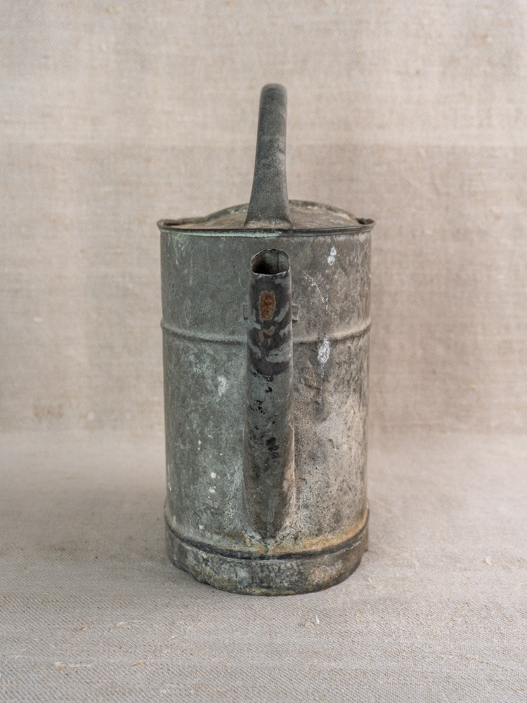 Antique French zinc watering can with curved handle and short spout, weathered finish on neutral fabric.