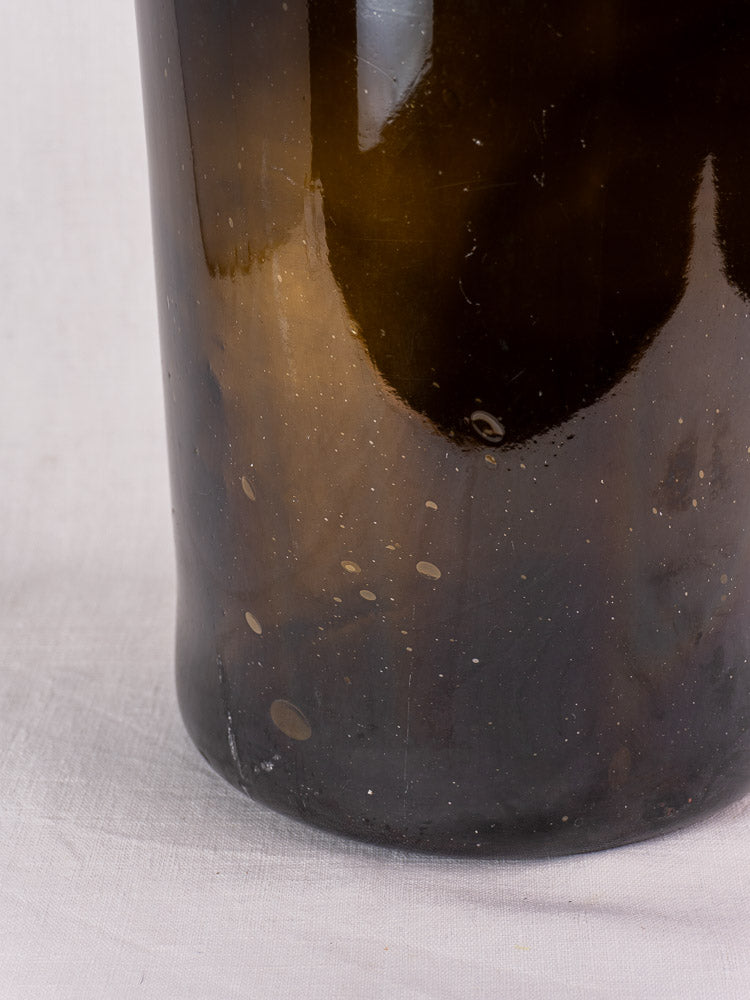 Antique French demijohn in dark olive hand-blown glass with original chain, cork, and air bubbles on white fabric background.