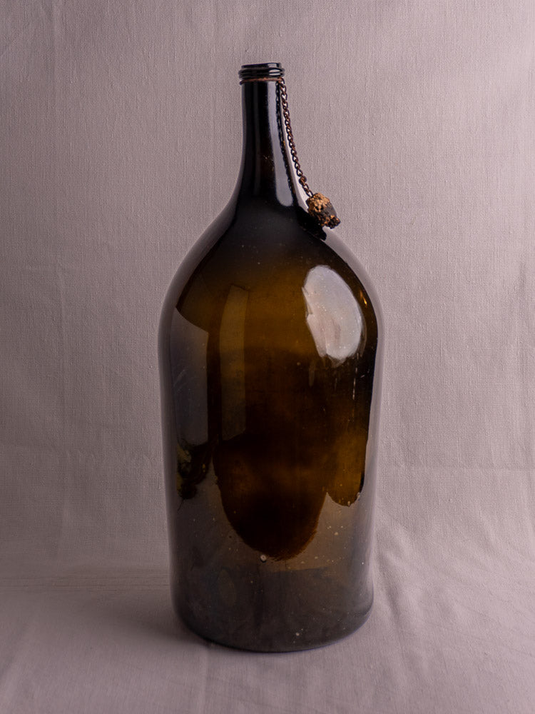 Antique French demijohn with dark olive hand-blown glass, narrow neck, chain, and cork on light fabric background.