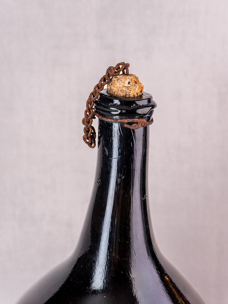 Antique French demijohn in dark olive hand-blown glass, original cork and metal chain on light background.