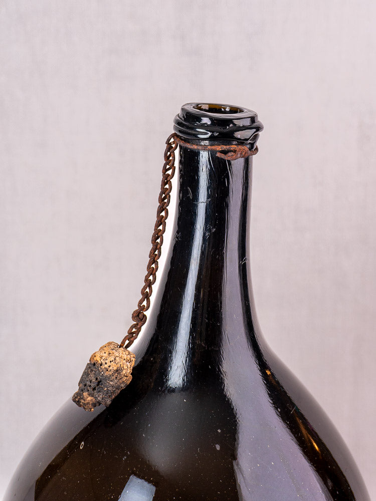 Antique French demijohn in dark olive hand-blown glass with original cork and chain, on a light background.