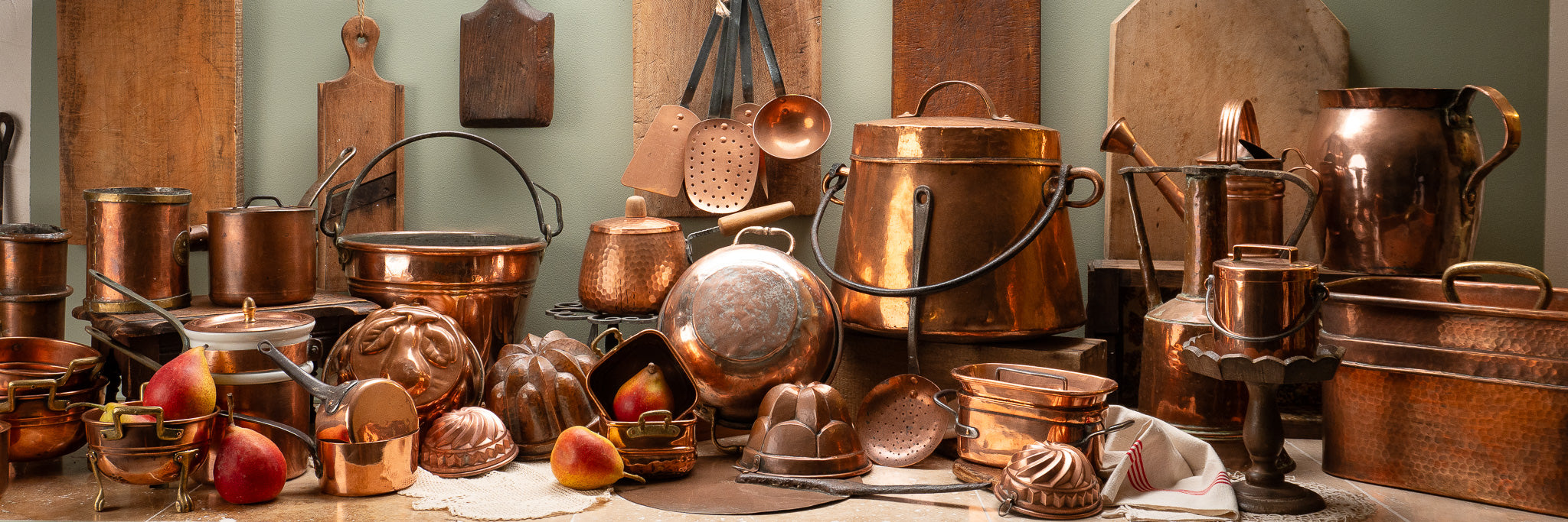 Vintage copper pots, pans, molds, and utensils with apples and pears on a counter; cutting boards in background.