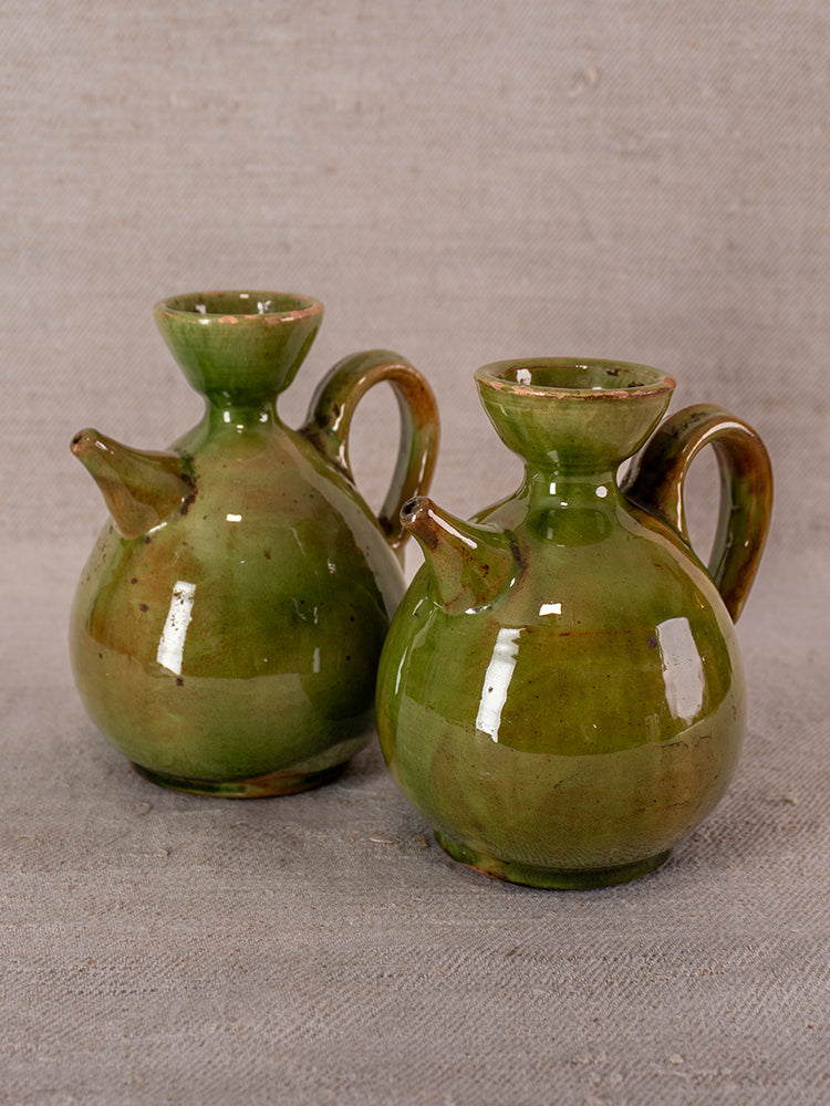 Pair of antique French green-glazed earthenware pitchers with round bodies and looped handles on grey fabric.