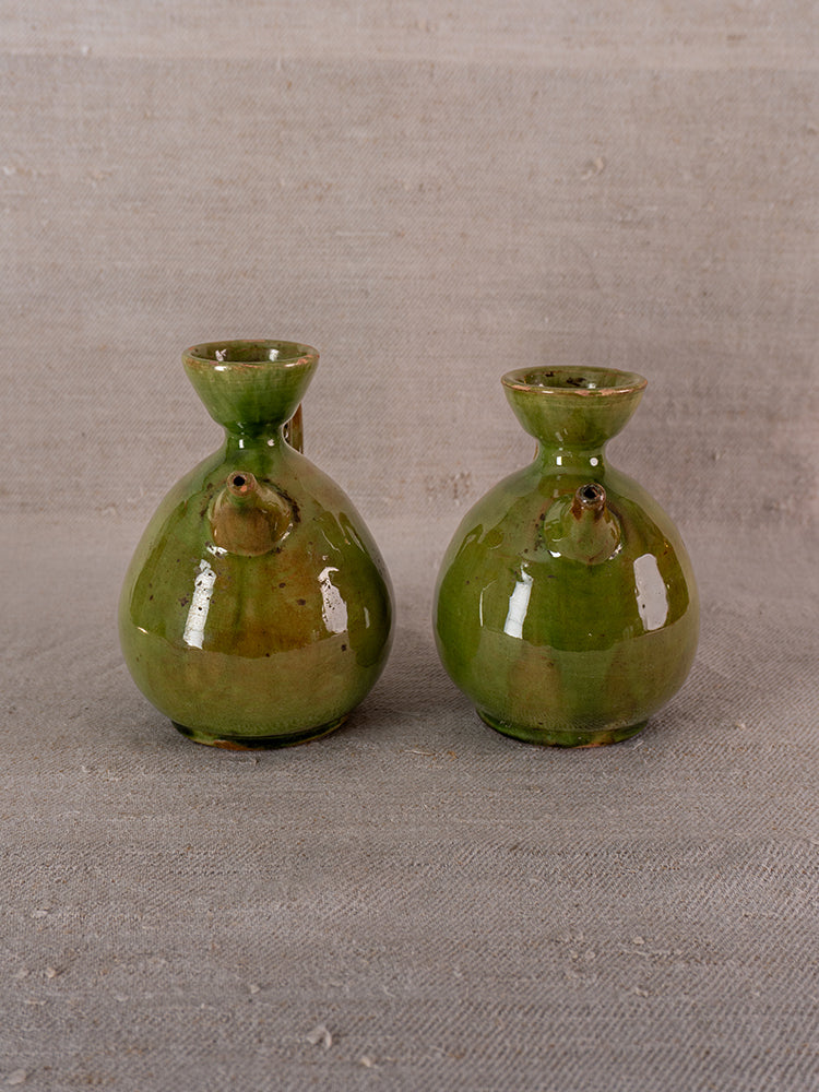 Pair of antique French green glazed earthenware pichets with round forms and front spouts on neutral fabric.
