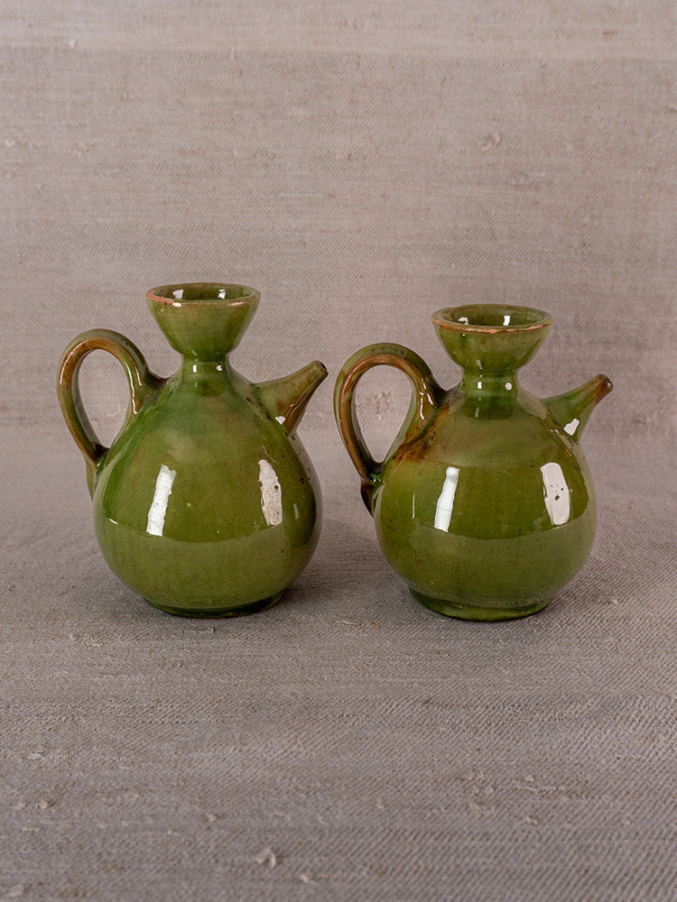 Pair of antique French green glazed earthenware oil and vinegar pitchers on gray textured fabric.